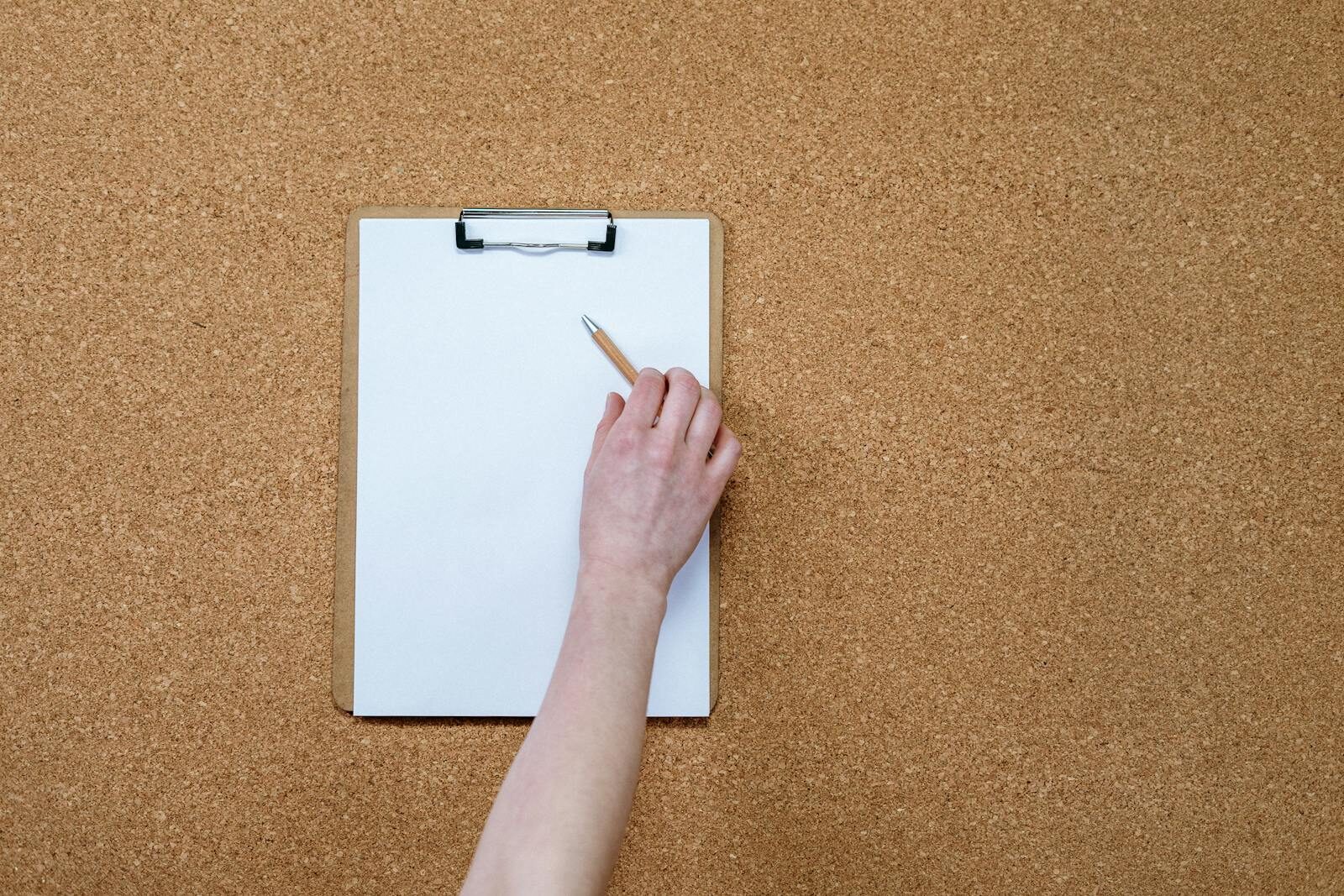 Close-up of a hand writing on a clipboard against a corkboard background.